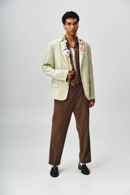 Genç bir Afrikalı Amerikalı adam, gri bir zemin üzerinde, çiçeklerle süslenmiş, kendine güvenen bej bir ceket giyiyor..