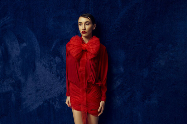 Молодая женщина с короткими волосами позирует в красном платье с большим бантом на синем фоне.