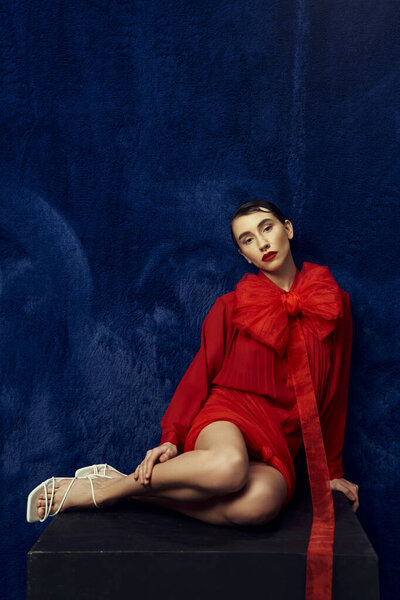 Молодая женщина с короткими волосами позирует в красном платье с большим бантом на синем фоне.