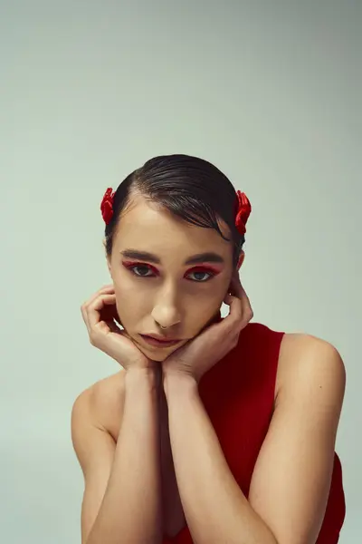 stock image Young woman with short, dark hair poses against a grey backdrop, looking thoughtful with hands on chin.