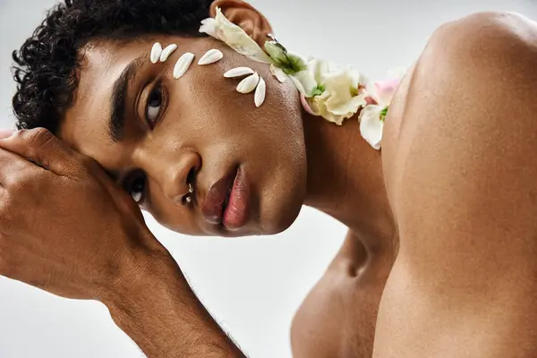 stock image A young, muscular African American man with flowers on his face and body poses against a grey backdrop, showcasing beauty and diversity.