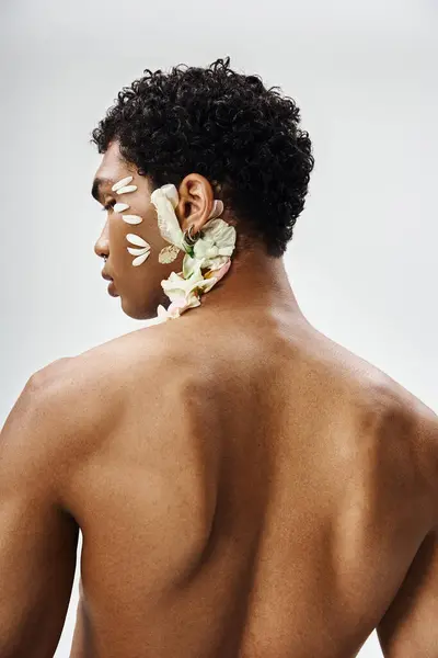 stock image A young, muscular African American man poses against a grey background, his face and body decorated with delicate white flower petals.