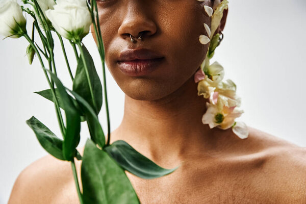 A close-up portrait of a young Black man with white flowers adorning his face and body against a gray background.