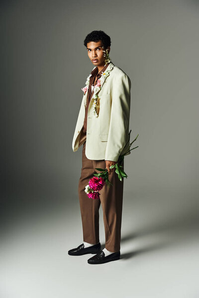 A young African American man with flowers adorning his neck and face poses against a grey background in a stylish blazer.