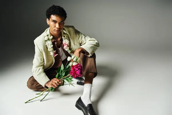 stock image A young African American man wearing a stylish blazer sits on a grey background with flowers on his neck