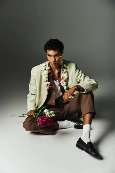 stock image A young Black man sits on grey background, with flowers around neck and face, wearing stylish blazer and brown pants.