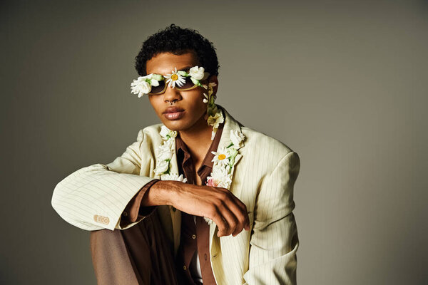 Молодой афроамериканец позирует в стильном блейзере, украшенном цветами на шее и лице, на сером фоне.