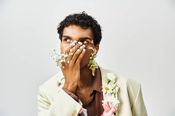 Молодой афроамериканец позирует с цветами, украшающими его шею и лицо на сером фоне, демонстрируя красоту и индивидуальность..
