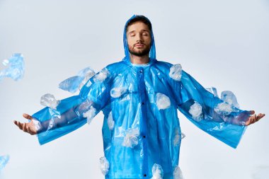 Kolları açık duran bir adam, plastik torbalarla süslenmiş mavi bir yağmurluk giyiyor..