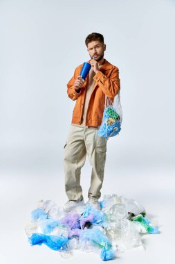A man stands on a large pile of plastic waste, holding a blue cup and a mesh bag. clipart