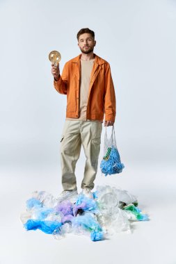 Adam plastik atık yığınının üzerinde duruyor, bir elinde ampul, diğer elinde ağ torbası..