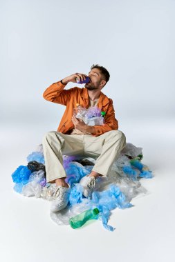 Plastik atık deniziyle çevrili bir adam oturur ve bir kutudan su içer..