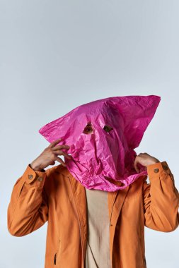 Parlak turuncu ceketli bir adam pembe plastik bir çantayla poz veriyor..