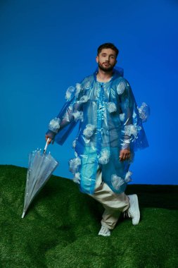 Plastikten yapılmış yağmurluk giyen bir adam elinde şemsiyeyle yeşil bir tepede duruyor..