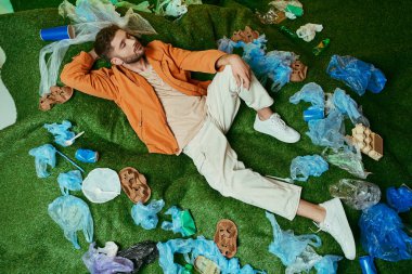 Yeşil bir çimenlikte yatan bir adam, plastik torbalar, şişeler ve diğer atıklarla çevrili..