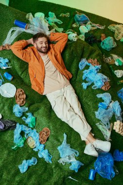 Plastik döküntülerle çevrili yeşil çimlerin üzerinde rahatça uzanan bir adam..
