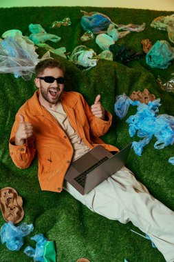 Yeşil çimenlikteki güneş gözlüklü adam, dizüstü bilgisayarı ve baş parmağı yukarıda, plastik atıkla çevrili..