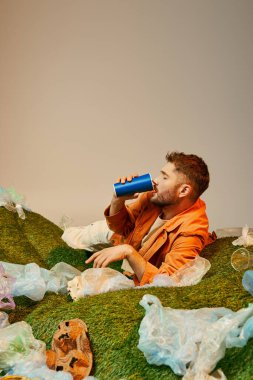 Plastik atık yığınında oturan bir adam mavi bardaktan içiyor..