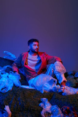 Bir adam, mavi ve kırmızı ışıklarla aydınlatılmış plastik torbalar ve molozların arasında oturur..
