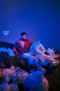 Genç bir adam, mavi ve kırmızı ışıkla aydınlatılmış plastik torbalar denizinin ortasında oturuyor..