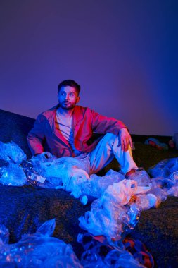 Plastik torbalar ve şişelerle çevrili, mavi ve mor ışıklarla aydınlatılmış bir adam oturuyor..
