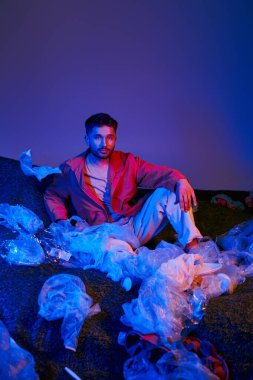 A man sits surrounded by plastic bags, bathed in blue and red light. clipart