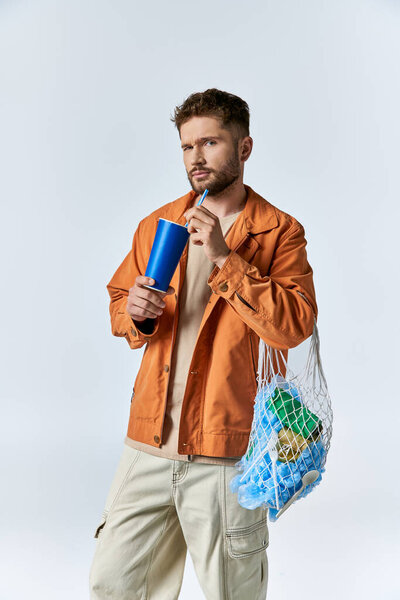 A man in a rust-colored jacket holds a blue cup and a white mesh bag filled with plastic.