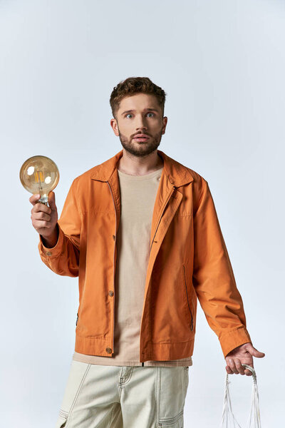 A man in an orange jacket holds a lightbulb and stares intently.