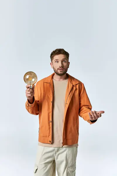 stock image A man in an orange jacket holds a lightbulb and stares upwards, lost in thought.