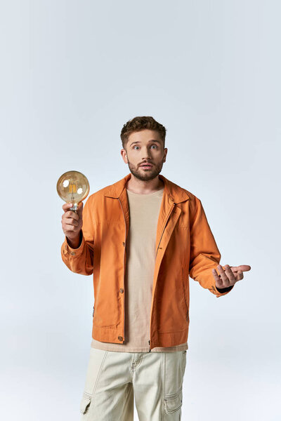 A man in an orange jacket holds a lightbulb and stares upwards, lost in thought.