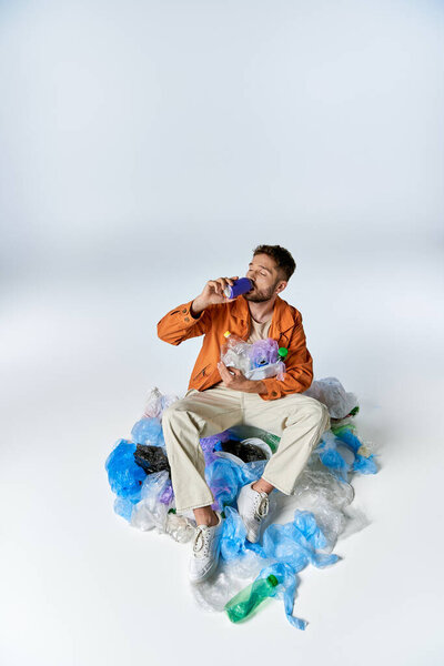 A man sits surrounded by plastic waste, drinking from a bottle.