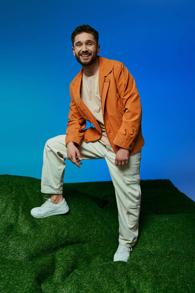 A handsome man in an orange jacket smiles on a green hill.