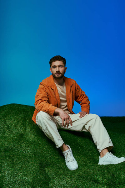 A man in an orange jacket sits on a green hill with a blue background.
