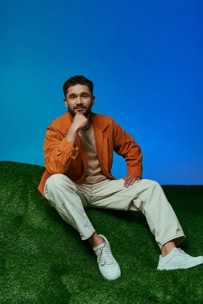 stock image A man in an orange jacket contemplates while sitting on artificial grass.