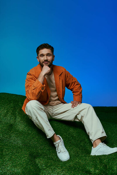 A man in an orange jacket contemplates while sitting on artificial grass.