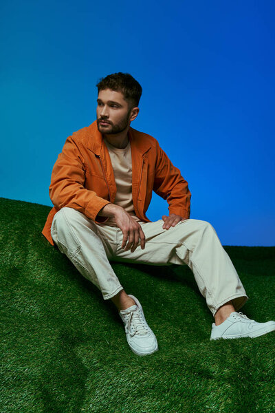 A young man sits on a patch of green grass, wearing a bright orange jacket.