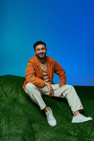 A man in an orange jacket and white pants sits on a patch of green grass.