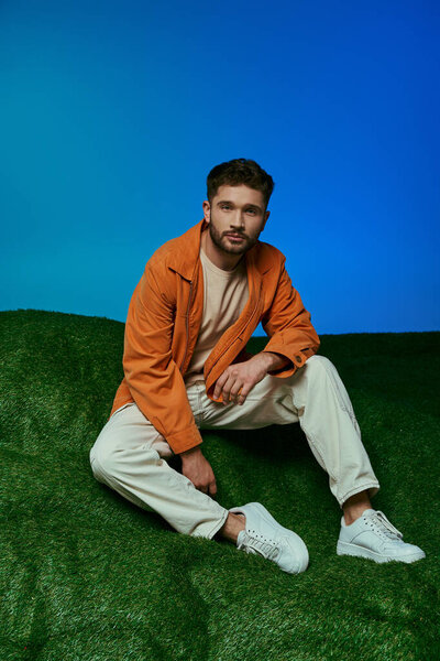 A young man wearing an orange jacket sits on artificial grass in front of a blue background.
