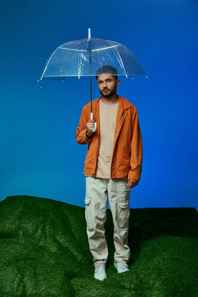 A man in an orange jacket stands on a green hill with a clear umbrella.