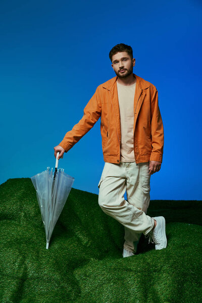 A man stands on a patch of artificial grass holding a clear plastic umbrella.