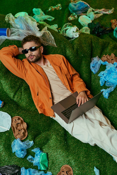 A man lies on fake grass, surrounded by plastic waste, using a laptop.