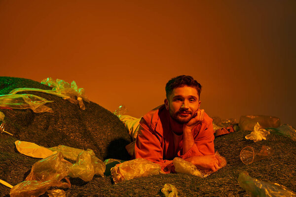 A man lies in a bed of plastic, surrounded by discarded items, staring up at the orange-toned sky.