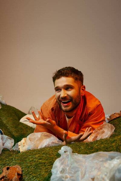 A man in an orange shirt laughs while lying on a bed of green grass, surrounded by plastic bags.