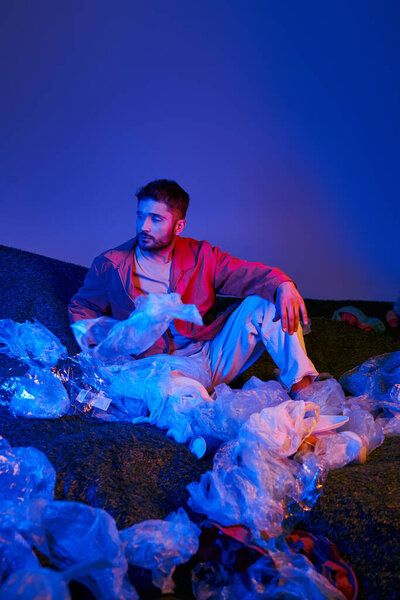Человек сидит в окружении моря пластиковых пакетов, купается в синем и красном свете.