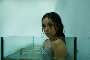 A beautiful young woman with wet hair, wearing a shimmering top, emerges from a glass tank filled with water. clipart