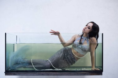 Deniz kızı gibi giyinmiş uzun, siyah saçlı genç bir kadın su dolu büyük bir cam tankın içinde yatıyor..