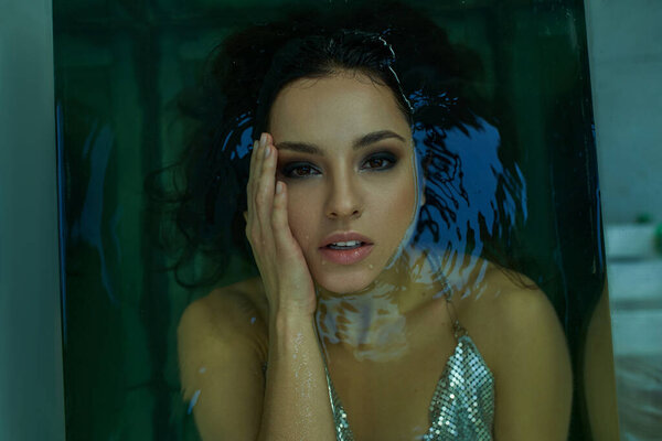 A young woman with wet hair, submerged in a blue-green pool, gazes intently through the water, her face illuminated by a soft light.