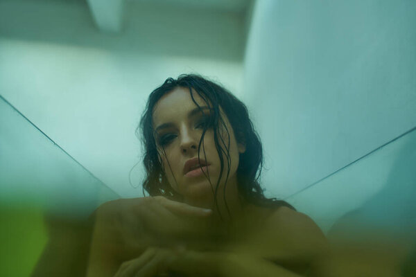 A young woman with wet hair emerges from the water, her gaze piercing through the glass.