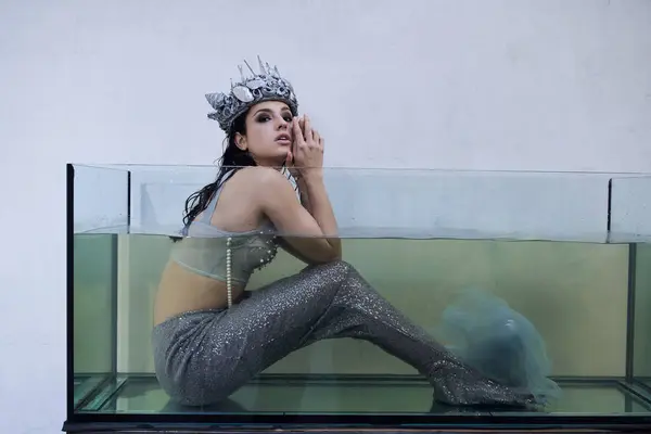 stock image A young woman wearing a crown and mermaid tail sits in a glass tank filled with water, gazing dreamily ahead.