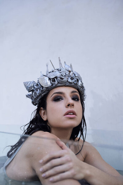 A young woman with dark hair, wearing a crown of seashells, sits submerged in water.